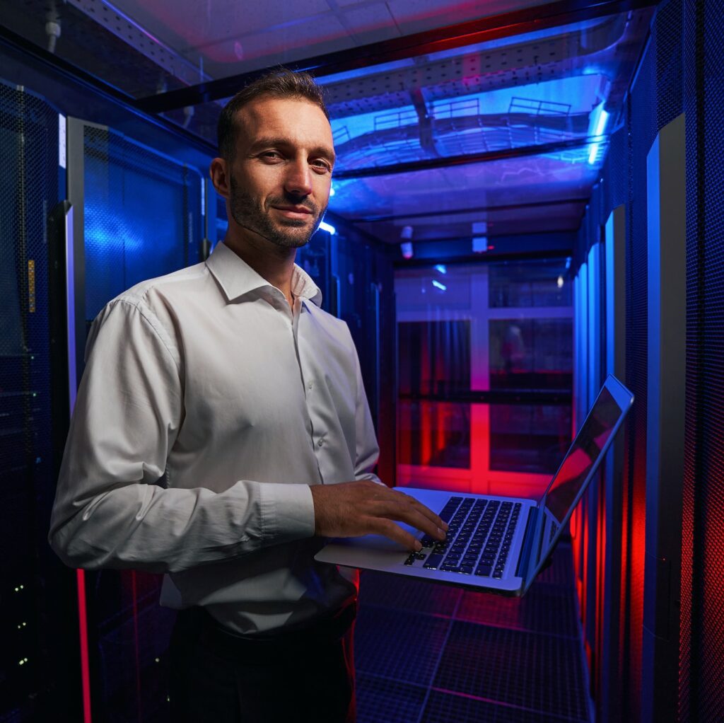 Bearded smiling system administrator is working with server rack and telecommunication equipments in